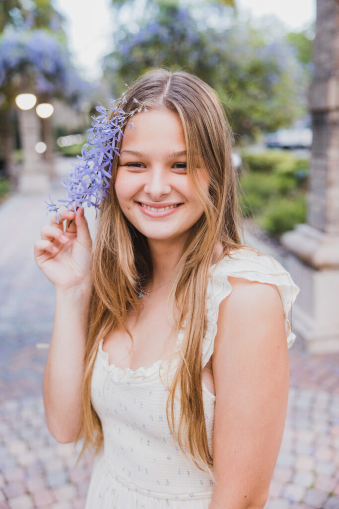 Image of a high school senior posing in a natural setting with a relaxed smile and a casual outfit, smiling with purple flowers. 