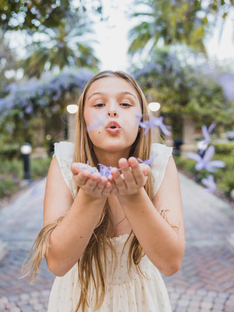 Image of a high school senior posing in a natural setting with a relaxed smile and a casual outfit, blowing purple flowers. 