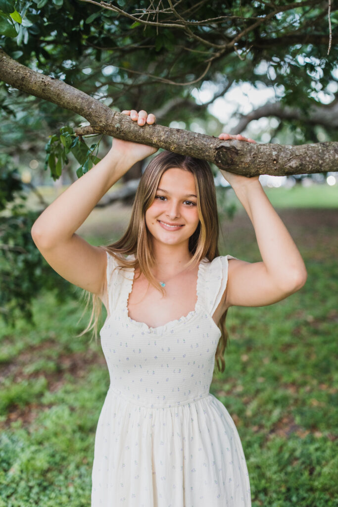 Image of a high school senior posing in a natural setting with a relaxed smile and a casual outfit, standing near a tree. 