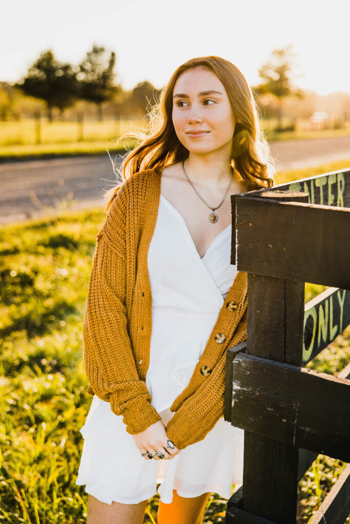 Image of a high school senior posing in a natural setting with a relaxed smile and a casual outfit, standing against a fence with the sun setting behind. 