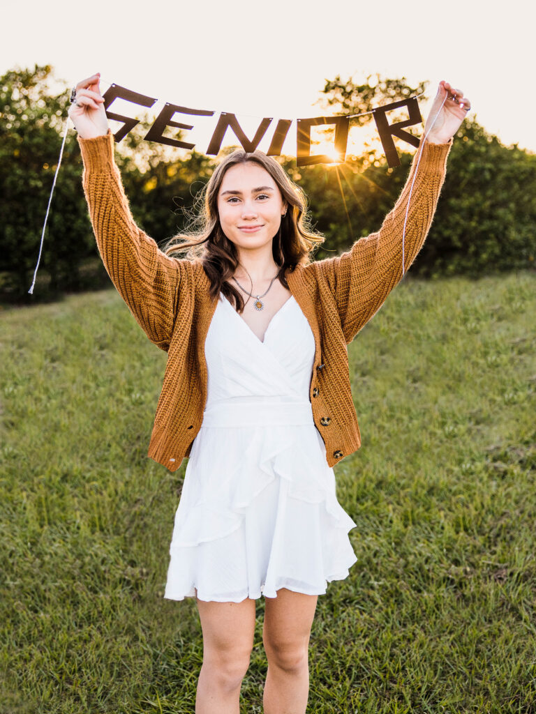 Image of a high school senior posing in a natural setting with a relaxed smile and a casual outfit, holding a Senior sign in the sunset.