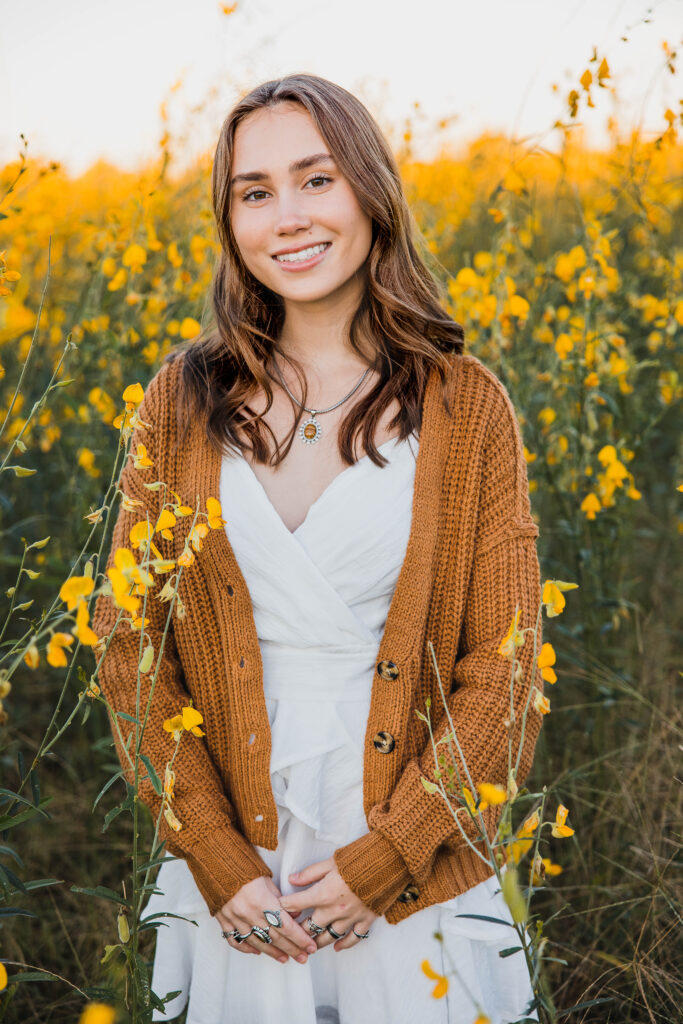 Image of a high school senior posing in a natural setting with a relaxed smile and a casual outfit, standing in a yellow field of flowers. 