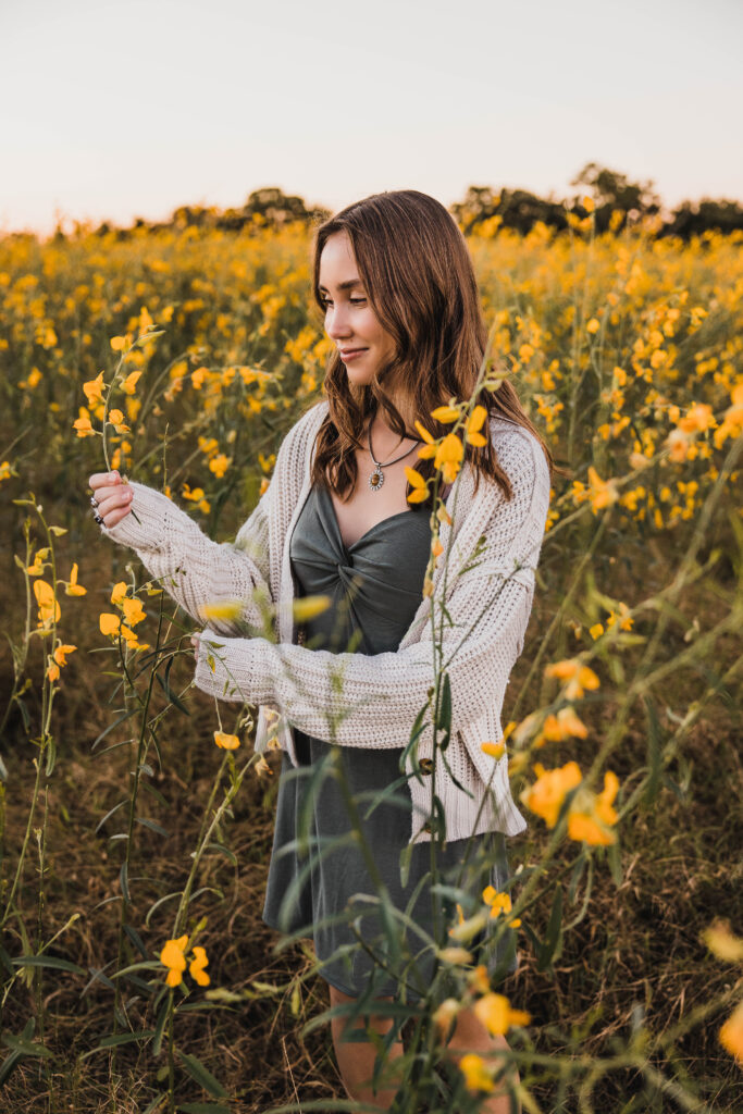Image of a high school senior posing in a natural setting with a relaxed smile and a casual outfit, standing in a yellow field of flowers. 