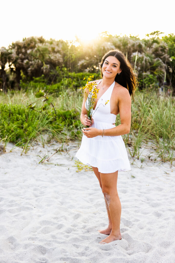 Image of a high school senior posing in a natural setting with a relaxed smile and a casual outfit, holding yellow flowers. 