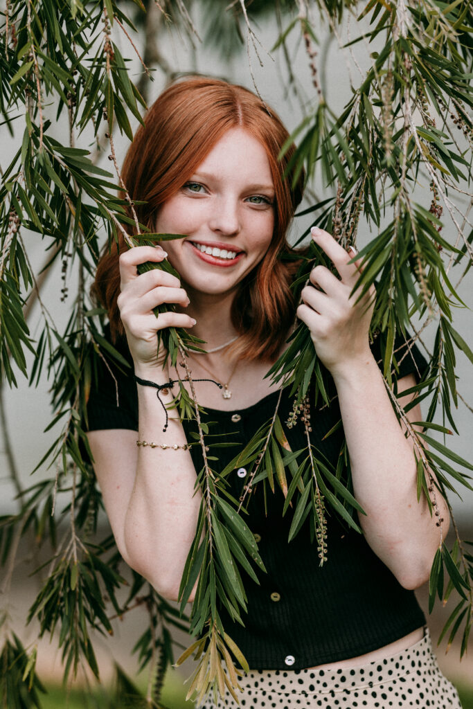 Image of a high school senior posing in a natural setting with a relaxed smile and a casual outfit, holding onto  tree branches. 