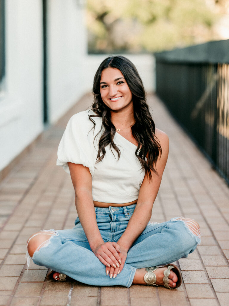 Image of a high school senior posing in a natural setting with a relaxed smile and a casual outfit, setting on a tiled floor. 