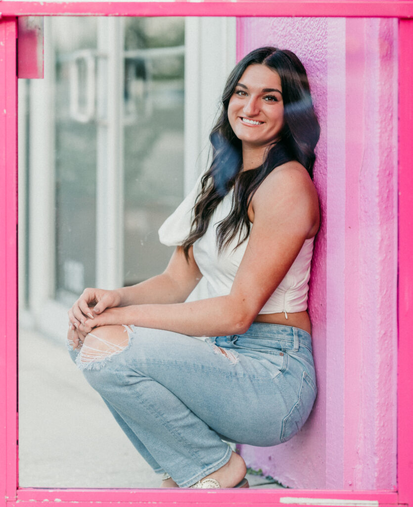 Image of a high school senior posing in a natural setting with a relaxed smile and a casual outfit, kneeling by a pink door. 
