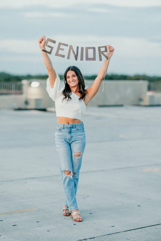 Image of a high school senior posing in a natural setting with a relaxed smile and a casual outfit, holding a senior sign on a parking garage. 