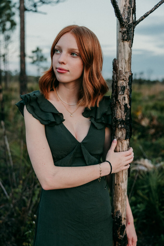 Image of a high school senior posing in a natural setting with a relaxed smile and a casual outfit, standing holding onto a tree. 