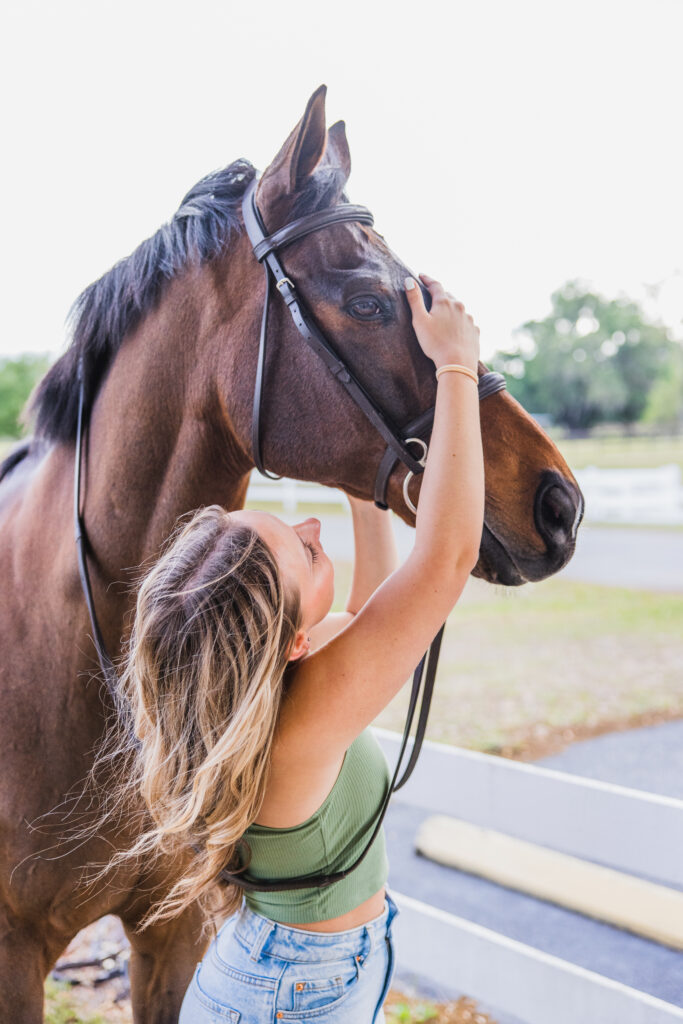 Image of a high school senior posing in a natural setting with a relaxed smile and a casual outfit, attending to her horse. 