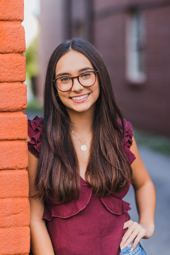 Image of a high school senior posing in a natural setting with a relaxed smile and a casual outfit, leaning on a brick wall.