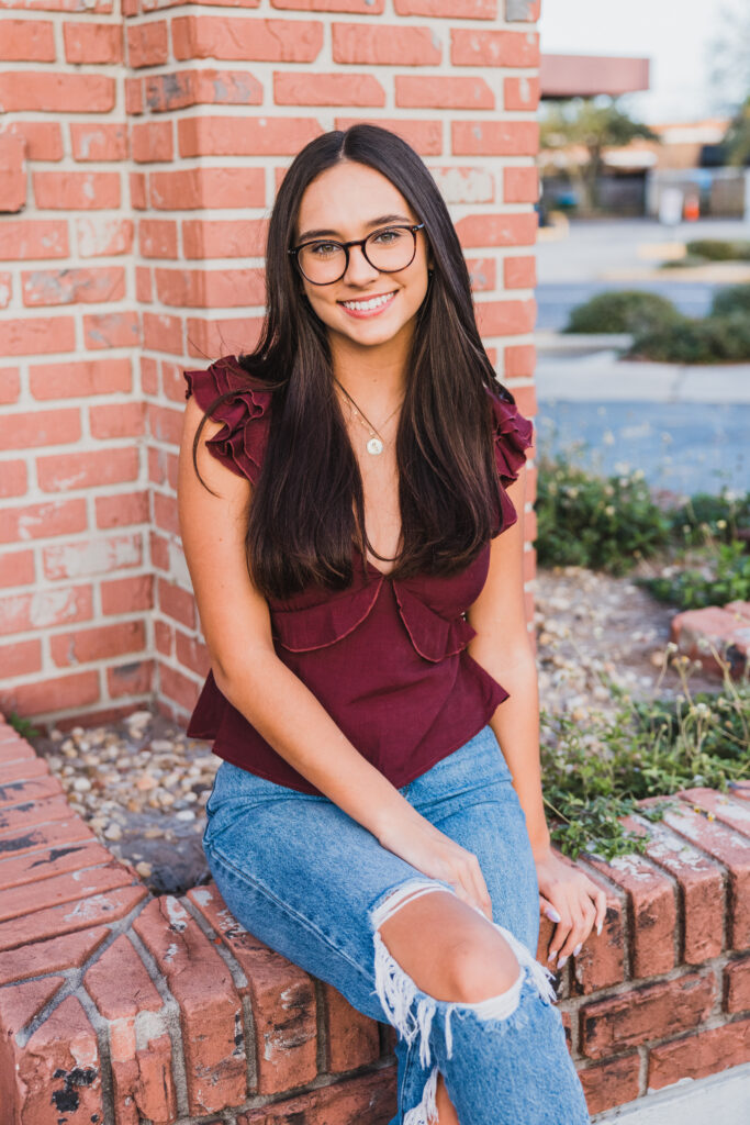 Image of a high school senior posing in a natural setting with a relaxed smile and a casual outfit, setting near a brick wall. 