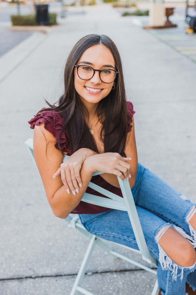 Image of a high school senior posing in a natural setting with a relaxed smile and a casual outfit, setting in a chair on a sidewalk.
