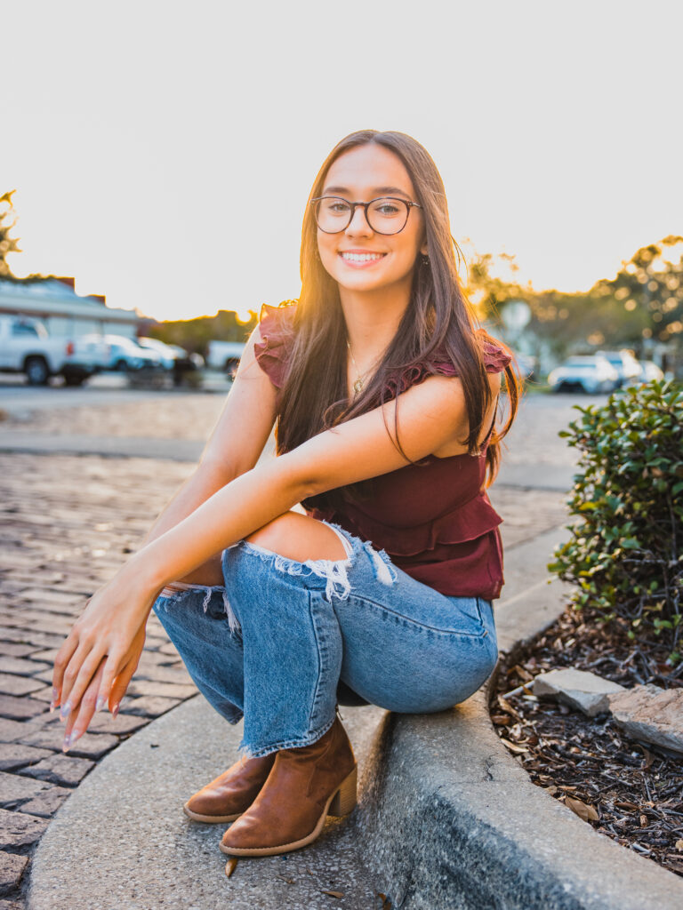Image of a high school senior posing in a natural setting with a relaxed smile and a casual outfit, setting on a street with a sunset.